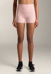 Ružové šortky s vysokým pásom vyrobené z hladkého, elastického materiálu, s obtiahnutým strihom a bezšvovým dizajnom. Skombinované s čiernymi teniskami.