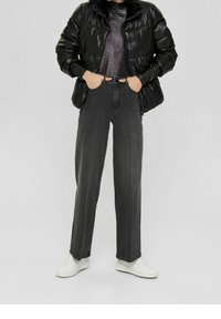 Schwarze gepolsterte Jacke mit hohem Kragen, getragen über einem dunklen strukturierten Oberteil, kombiniert mit gerade geschnittenen dunkelgrauen Jeans und weißen Sneakers.