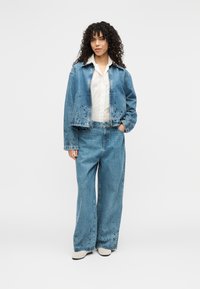 Τζιν μπουφάν και παντελόνι με πλατύ μπατζάκι σε ανοιχτό μπλε με διακοσμητικές λεπτομέρειες. Λευκό πουκάμισο με κουμπιά από κάτω. Τα παπούτσια είναι εκρού.