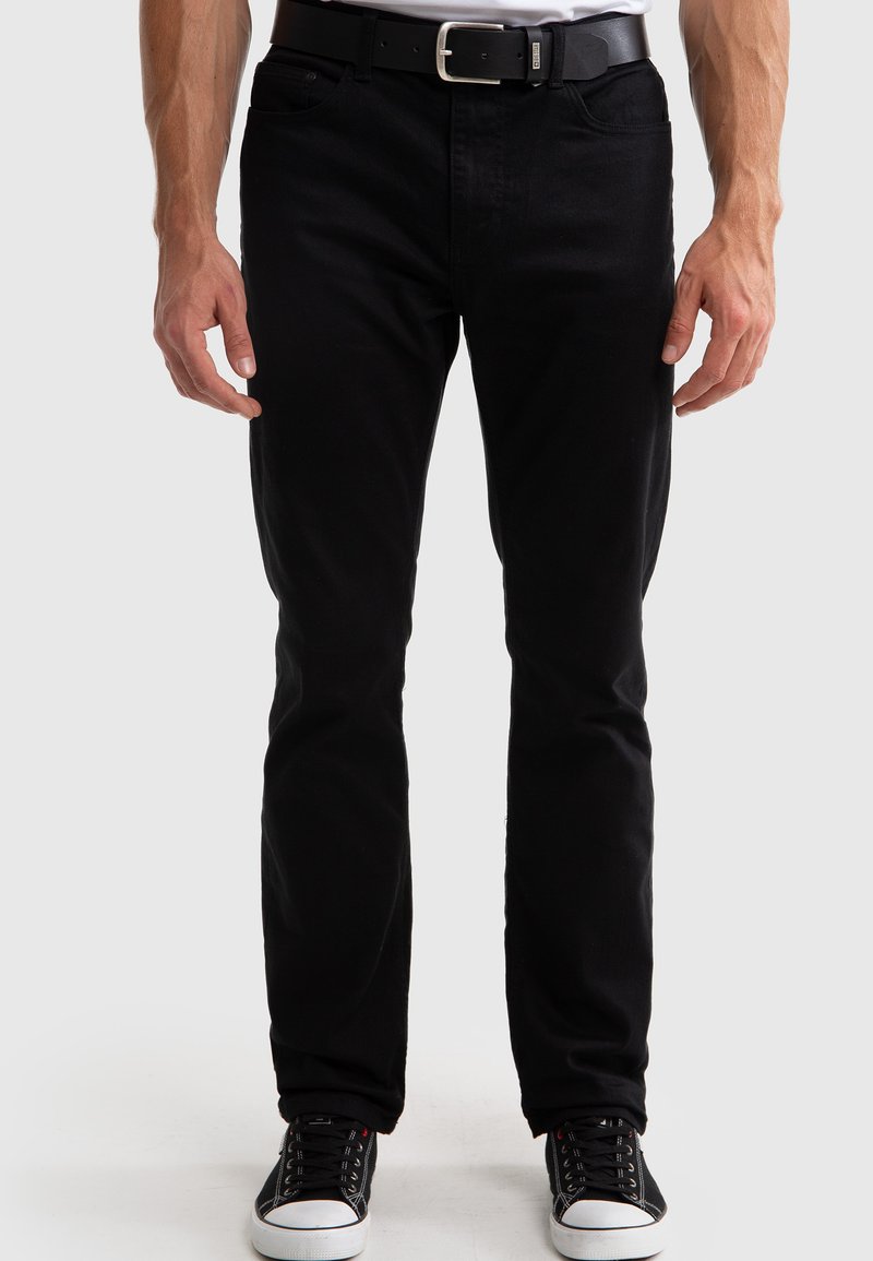Jeans de mezclilla negros con un corte slim, diseño de cinco bolsillos y detalles de costura sutiles; llevados con un cinturón negro y zapatillas negras.