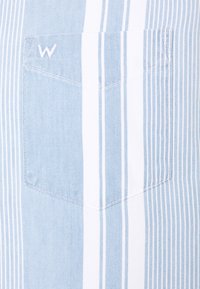 Modrá a bílá látka s vertikálními pruhy má malou náprsní kapsu s vyšitým písmenem "W". Textura je hladká a lehká.