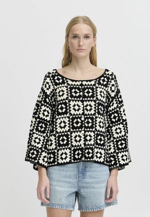Pullover a righe in bianco e nero con scollo ampio e maniche a campana, caratterizzato da un design geometrico e un tessuto textured. Abbinato a pantaloncini di jeans.