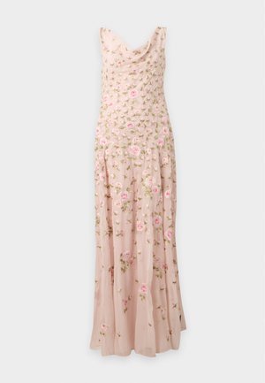 Femme aux longs cheveux foncés portant une robe maxi rose sans manches brodée de fleurs et des chaussures en satin pointues, debout avec les mains dans le dos.