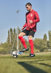 Cămașă roșie cu mâneci lungi și logo, șorturi negre, șosete roșii și ghete de fotbal negre, driblând o minge de fotbal alb-negru pe un teren de iarbă.