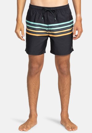 Man in zwarte zwembroek met horizontale turquoise, gele en oranje strepen, die blootsvoets op een witte achtergrond staat.