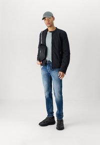 Crna bomber jakna, siva majica, plave isfucene traperice, crne tenisice i siva kapa, drži crnu torbu s trakama s logotipom.