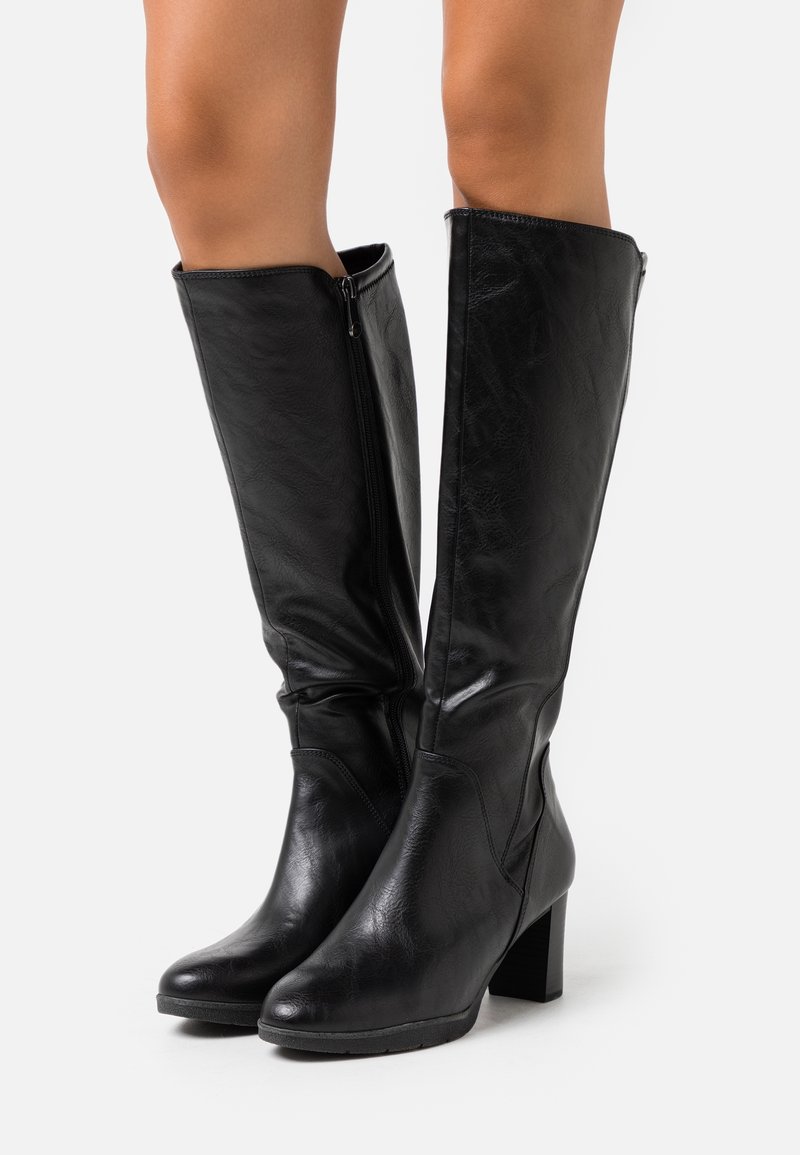 Paire de bottes en cuir noires montant jusqu'aux genoux avec fermetures éclair latérales et talons blocs portées par une personne sur fond blanc.