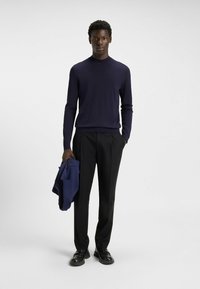 Maglione lungo blu in tessuto a maglia fine, abbinato a pantaloni neri su misura. Modello che tiene in mano una giacca blu, in piedi su uno sfondo bianco.