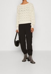 Krämfärgad stickad tröja med öppet mönster och ribbad fåll, ihop med svarta cargo-byxor och grå klackade ankelskooter. Svart handväska.