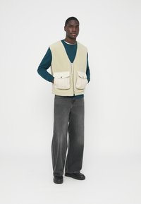 Chaleco de forro polar en verde pálido con dos bolsillos delanteros beige, llevado sobre una sudadera azul verdoso. Combinado con vaqueros anchos grises y zapatos negros.