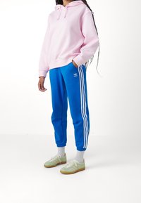 Felpa con cappuccio rosa chiaro, pantaloni jogger blu con strisce bianche e logo, e scarpe da ginnastica verdi con accenti beige.