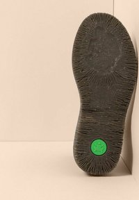 Suela de zapato negro desgastada con una fina textura acanalada y un logo circular verde que presenta una rana cerca del talón, posicionada contra paredes beige.