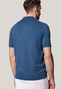 Niebieska koszulka polo z krótkim rękawem z miękkiego dzianiny. Posiada prążkowany kołnierzyk i dół, z bezszwowym projektem ramion, pokazana od tyłu.