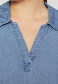 Lyseblå denimskjorte med V-hals, med en foldet krave, synlige syninger og en blød textile texture.