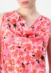 Blomsterert top uten ermer i rosa med livlige oransje, svarte og brune blomster. Har en drapert halslinning, glatt tekstur og lett stoff.