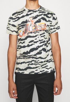 Hombre con camiseta negra y blanca de estampado animal con logo naranja "Just Cavalli" y pantalones negros, posando frente a un fondo liso.