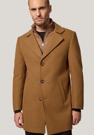 Mężczyzna w brązowym wełnianym płaszczu nałożył naquiltowany jasnobrązowy kurtkę z zamkiem z przodu, stoi na tle jednolitego jasnoszarego tła.