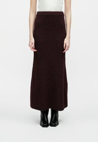 Vrouw in een lange, donkerpaarse gebreide rok en zwarte enkellaarsjes, staande tegen een effen witte achtergrond.