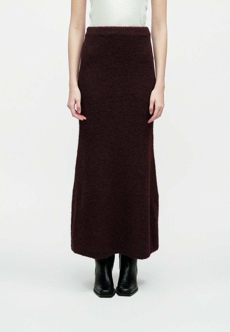 Vrouw in een lange, donkerpaarse gebreide rok en zwarte enkellaarsjes, staande tegen een effen witte achtergrond.