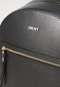 Fekete DKNY hátizsák texturált felülettel, arany cipzárral és logóval. Lekerekített tetejű forma, elülső cipzáras zsebével és minimalista dizájnnal.