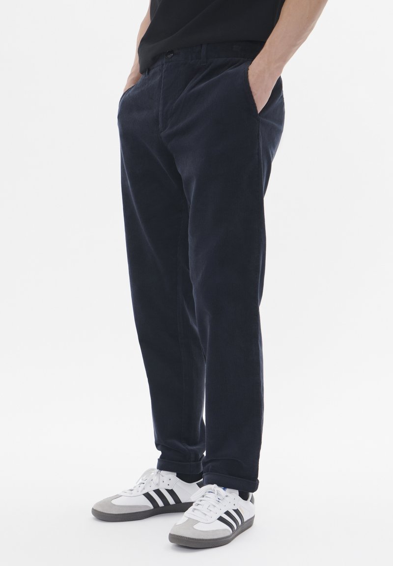 Homme portant un pantalon en velours côtelé bleu foncé et des baskets blanches à rayures noires, debout les mains dans les poches.
