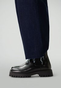 Bottines en cuir noir avec un bout rond, semelle épaisse noire et pannels élastiques sur les côtés. Portées avec un pantalon bleu foncé à l'ourlet effiloché.