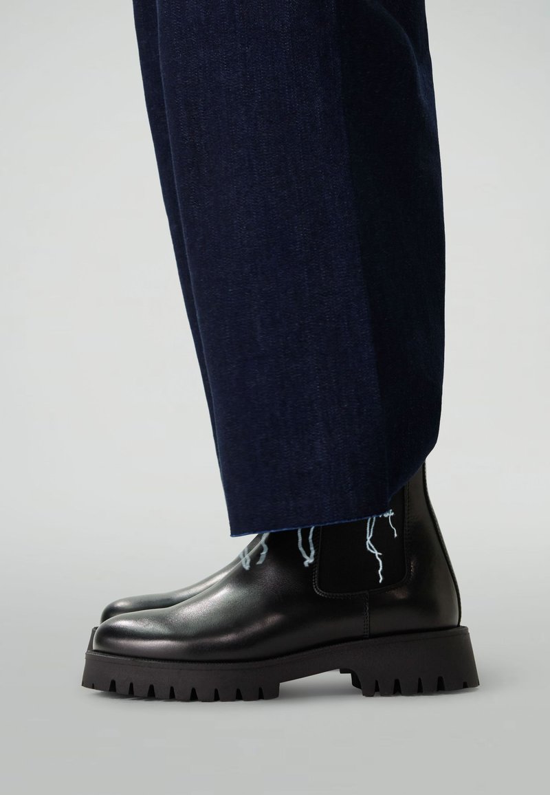 Bottines en cuir noir avec un bout rond, semelle épaisse noire et pannels élastiques sur les côtés. Portées avec un pantalon bleu foncé à l'ourlet effiloché.
