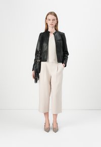 Giacca di pelle nera con colletto alto, top crema con texture, pantaloni beige chiaro a gamba larga e décolleté grigi a punta. Borsa a spalla con frange.