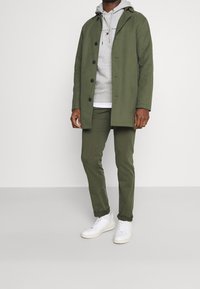 Olijfgroene jas met knopen, grijze hoodie, olijfgroene broek en witte sneakers. Heeft een capuchon en contrasterende texturen.