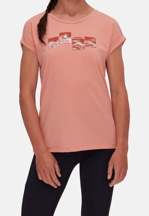Vrouw draagt een perzikkleurig T-shirt met korte mouwen met een berglandschap en de tekst "Mammut", gecombineerd met een zwarte broek.