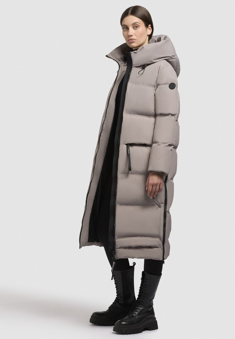 Long manteau rembourré gris clair avec un col haut, doté de poches zippées et d'une texture lisse. Porté sur une tenue noire et des bottes noires à lacets.