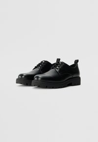 Zapatos de cuero negro con cordones, que presentan una puntera redondeada, suela de goma gruesa con estrías y una pestaña en forma de lazo en el talón para facilitar su uso.