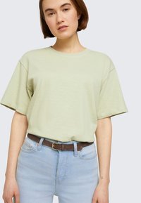 Šviesiai žalia oversize marškinėliai su trumpomis rankovėmis, pagaminti iš švelnios medžiagos, derinami su šviesiai mėlynomis aukšto liemens džinsais ir rudą diržu.