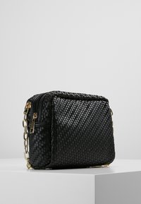 Sac en noir tressé de forme rectangulaire, avec un matériau texturé, une bandoulière en chaîne dorée et une fermeture éclair sur le dessus.