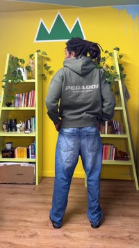 Green hoodie with "PEGADOR" logo on the back, paired with faded blue jeans, standing against a yellow wall and green shelving unit.