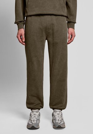 Person trägt olivgrüne Jogginghosen und weiße-graue Sportschuhe, steht vor einem schlichten hellgrauen Hintergrund.