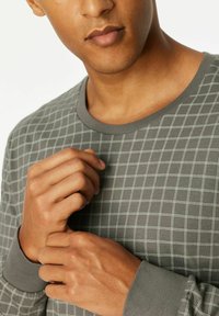 Grijze lange mouwen shirt met een licht ruitpatroon. Heeft een ronde hals en manchetten. Stof lijkt zacht met een gladde textuur. Handen die de stof vasthouden.