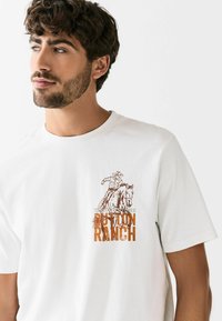 Valge puuvillane T-särk, millel on graafika cowboy'st hobusel, mille peal on oranž "Dutton Ranch" ja hall "Yellowstone". Lühikesed varrukad.