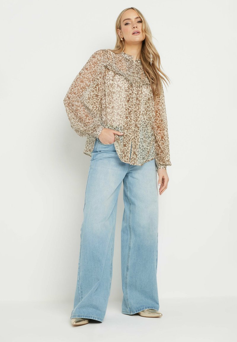 Durchscheinende Bluse mit Blumenmuster in warmen Tönen, kombiniert mit hellblauen, weit geschnittenen Jeans. Auffällige Puffärmel und runder Ausschnitt sorgen für Textur.