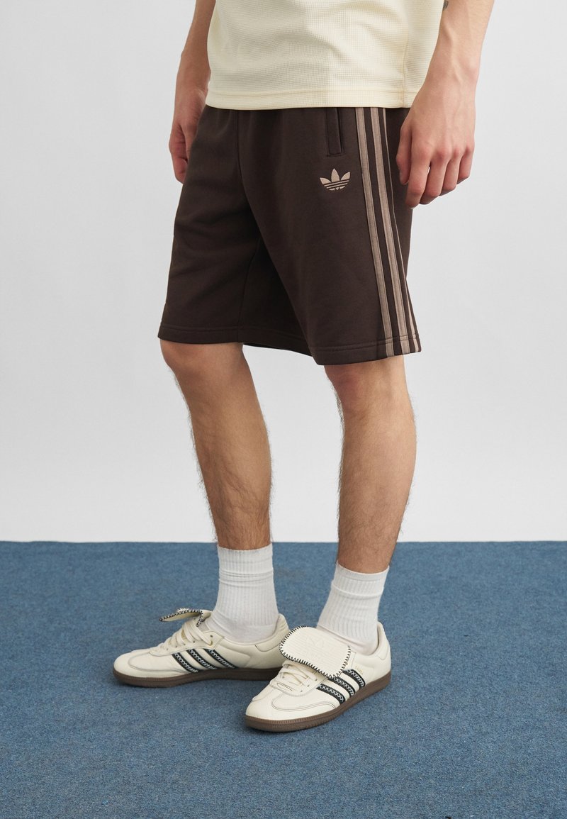 Mann trägt dunkelbraune Adidas-Shorts mit beigen Streifen und weiße Adidas-Sneakers mit schwarzen Streifen auf blauem Teppichboden.