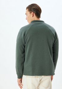 Sudadera verde oscuro con cuello y puños acanalados. Tejido suave, corte relajado y detalles en las costuras traseras. Se lleva con pantalones de color claro.
