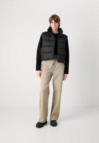 Colete puffer preto com gola alta, design acolchoado e botões de pressão, combinado com jeans de perna larga em bege claro e sapatos plataforma pretos.