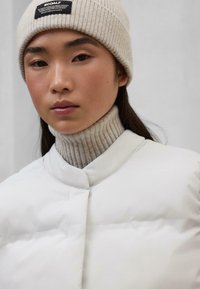 Chaqueta de plumas blanca con un alto cuello, acentuada por un cuello de tortuga de punto beige acanalado y un gorro de punto beige claro, que presenta un parche de logo.