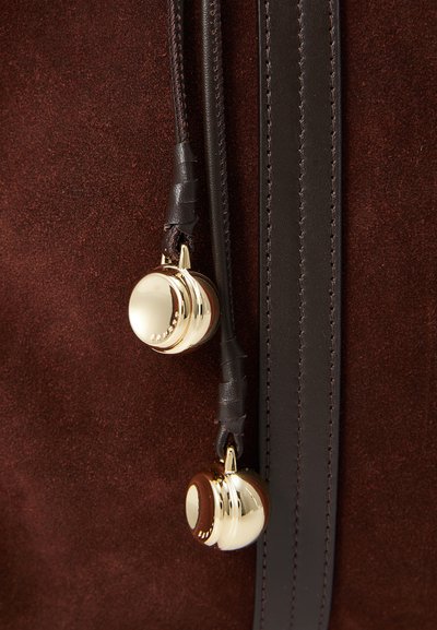 Détail d'un sac en suède marron avec quincaillerie dorée, des cloches décoratives, des accents en cuir noir et des coutures contrastées.