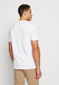 White short-sleeved t-shirt, round neckline, smooth cotton fabric, standard fit, worn with beige trousers, plain back view.