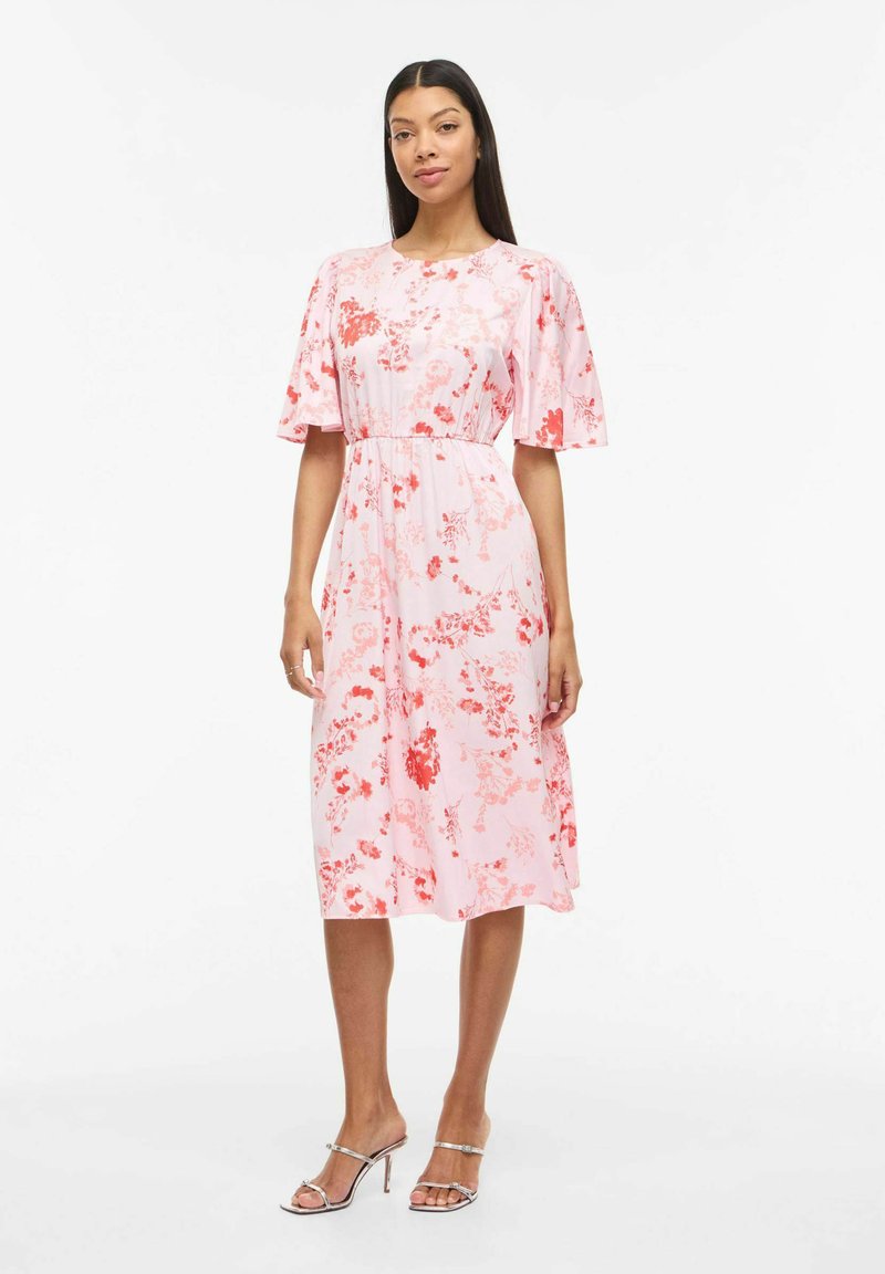 Femme debout portant une robe midi rose avec des motifs floraux rouges et des manches volantées, associée à des sandales argentées à talons hauts et à bouts ouverts.