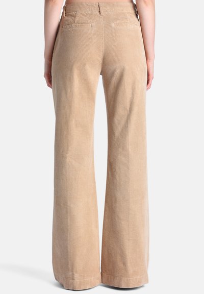 Personne portant un pantalon large en velours côtelé beige, vue de dos sur un fond clair uni.
