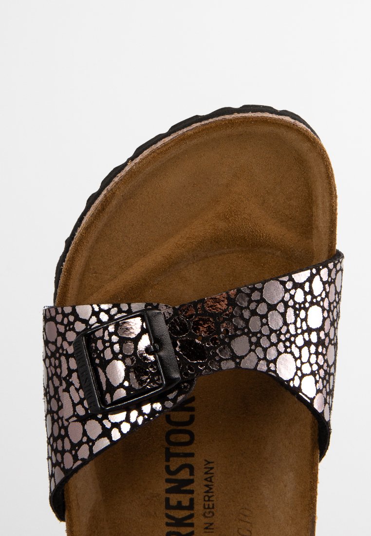 Schwarz und metallisch bronze Birkenstock-Sandale mit einem strukturierten Riemen, der ein kreisförmiges Muster aufweist, einer flachen, braunen Wildledersohle und einem verstellbaren Schnallenverschluss.