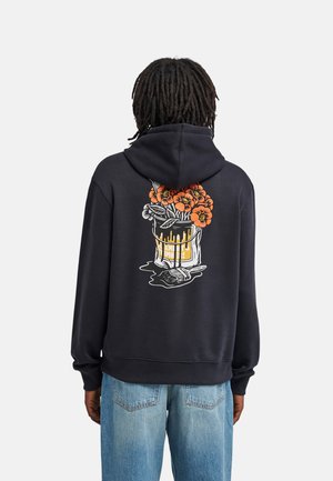 Personne aux dreadlocks foncés porte un sweat à capuche noir avec un motif de fleurs orange dans un seau de peinture qui dégouline dans le dos, associé à un jean bleu clair.