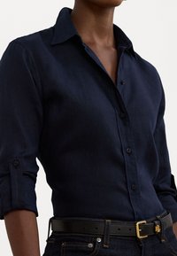 Marinblå långärmad skjorta med knappunderkrage, texturerat tyg och uppvikta ärmar, kombinerad med mörka jeans och ett svart skärp med en guldbuckle.